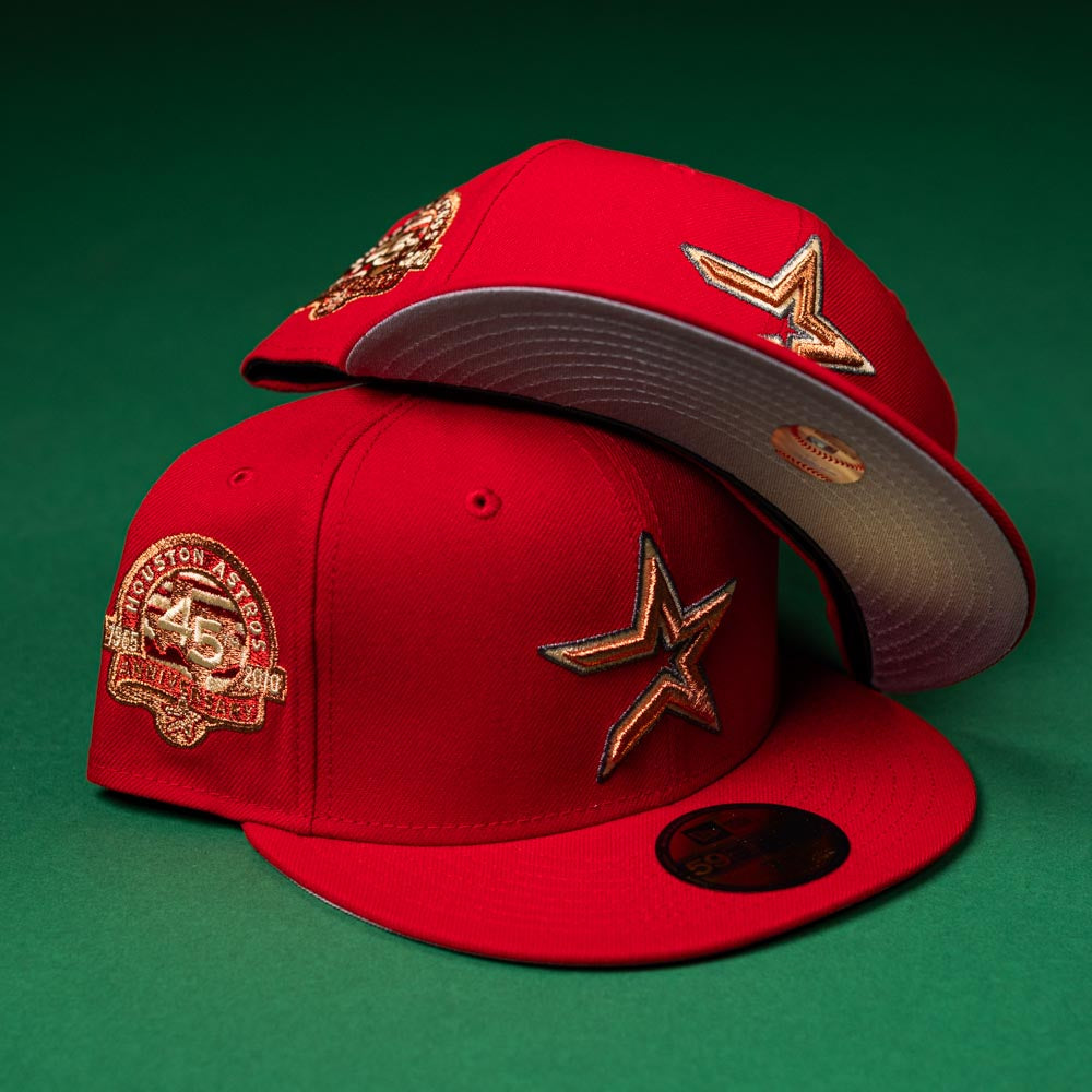 Two Rusty Screw OG Scarlet Copper Houston Astros New Era 5950 fitted hats are placed on a green surface; one is upright, while the other is upside down to display the underbrim in the signature Rusty Screw colorway. Both caps feature gold embroidered logos and anniversary patches.
