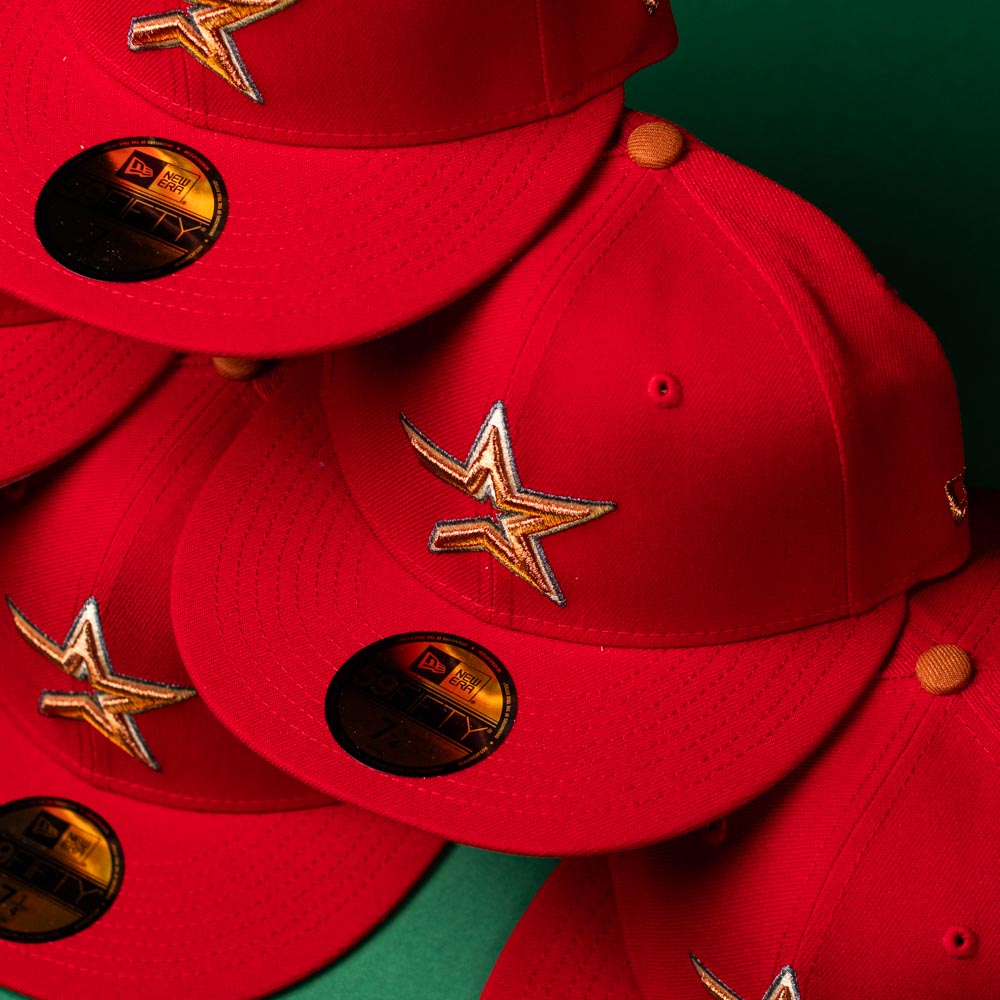 Multiple Rusty Screw OG Scarlet Copper Houston Astros New Era 5950 Fitted hats are arranged overlapping on a green surface in a close-up view. Each hat features a bright scarlet color, copper star logo, official stickers on the brim, and signature Astros detailing.