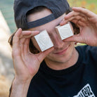 A person in a black shirt and backwards cap smiles while holding two beige rubber stamps from the SC Stamp Set Natural OS Unisex Santa Cruz over their eyes—one with a logo, the other with an engraved graphic design.