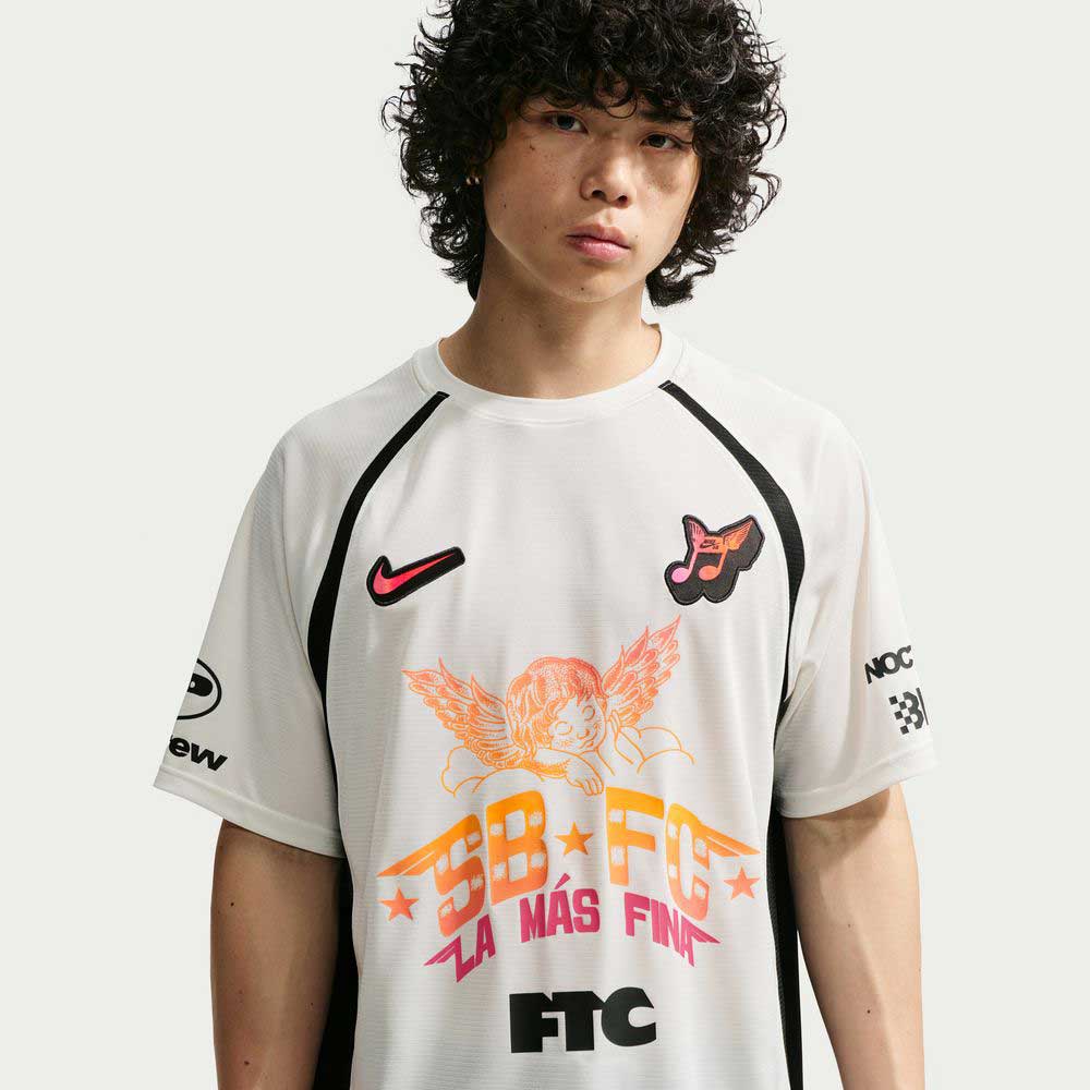 A young person with curly hair wears the Sail Black IR6760133 Skate Jersey from Nike SB Skateboarding, featuring colorful cherub and star graphics, plus Nike and FTC logos. The jersey is white polyester. Background is plain and light-colored.