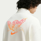 A person with curly hair wears the white Nike SB Skateboarding Sail IM5463133 1/2 Zip Fleece Crew, featuring a pink and orange sleeping angel illustration with wings and a Nike swoosh on the back; machine washable for easy care.