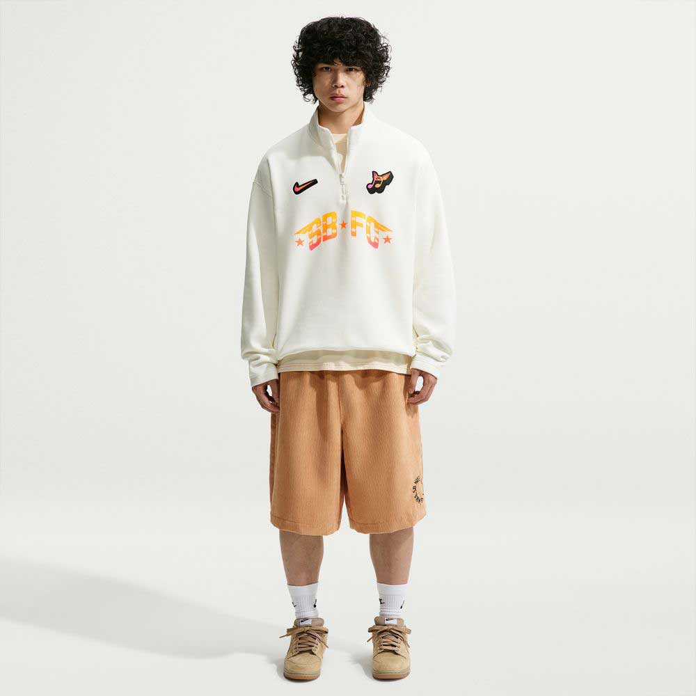 A young man with curly hair wears the Sail IM5463133 Skate 1/2 Zip Fleece Crew Nike SB Skateboarding, tan knee-length shorts, white socks, and tan sneakers, standing against a plain white background.