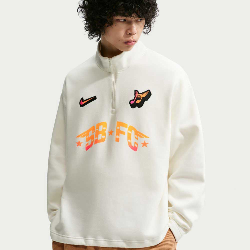 A curly-haired person wears the Nike SB Skateboarding Sail IM5463133 1/2 Zip Fleece Crew, a white imported sweatshirt with colorful patches—Nike swoosh, winged shoe, SB-FC stars—standing out against a plain light background. Machine washable.