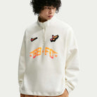 A curly-haired person wears the Nike SB Skateboarding Sail IM5463133 1/2 Zip Fleece Crew, a white imported sweatshirt with colorful patches—Nike swoosh, winged shoe, SB-FC stars—standing out against a plain light background. Machine washable.