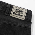 Close-up of Distressed Flat Black Relaxed Denim Jeans by Butter Goods, made from 100% cotton, featuring a beige “Butter” patch with globe graphic on the waistband.