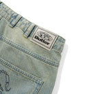 Scorpion Stitch Tinted Light Blue Denim Jeans by Butter Goods feature a baggy fit and a beige patch with a globe graphic and "Butter" in bold black above the back pocket. Shown on a white background.