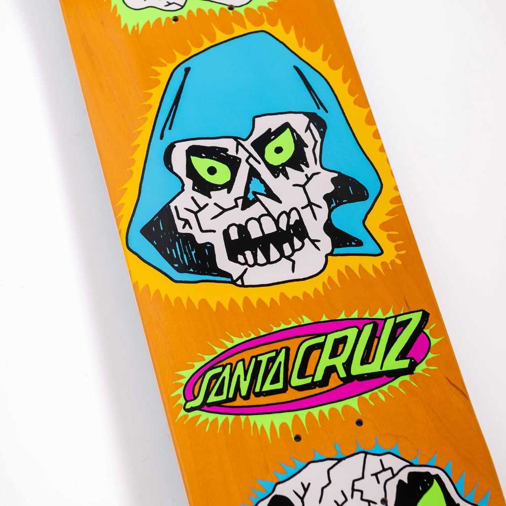 A close-up of the Scribble Head Feel Base 7.75in x 30.25in Santa Cruz Skateboards Deck shows a stylized skull with green eyes, a blue hood, and a colorful Santa Cruz logo on an orange background.
