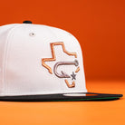 The Backlash Corpus Christi Hooks New Era 5950 Fitted Hat features a silver, black, and white design with an embroidered Texas outline, fishing hook, and star—resembling the iconic team cap—set against an orange background.