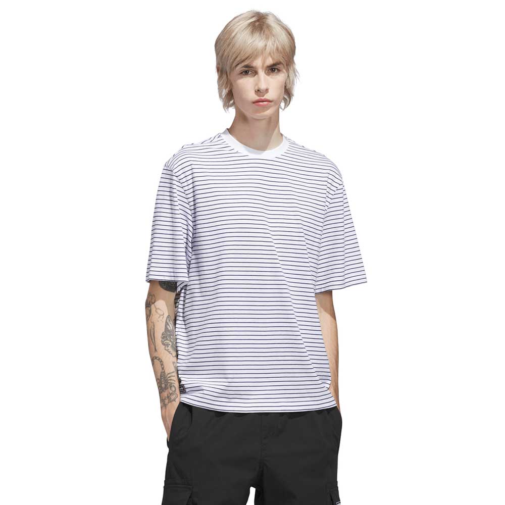 Person with light hair wearing an Adidas Skateboarding Skate Yarn Dye Tee in White/Dark Blue (KC6291) and black pants, standing with hands in pockets against a plain white background. Tattoos are visible on one arm.
