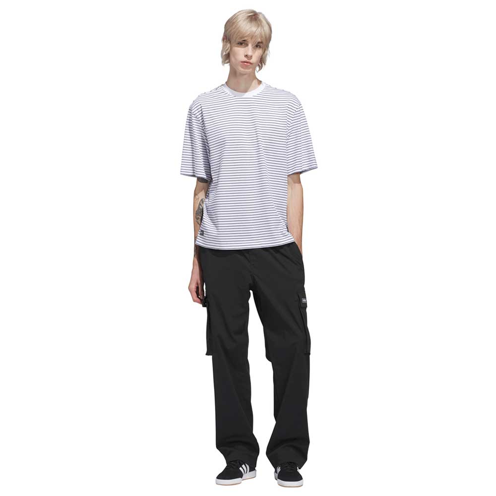 Person with short blond hair wearing the Skate Yarn Dye Tee White Dark Blue KC6291 by Adidas Skateboarding, loose black cargo pants, and black sneakers, standing against a white background.