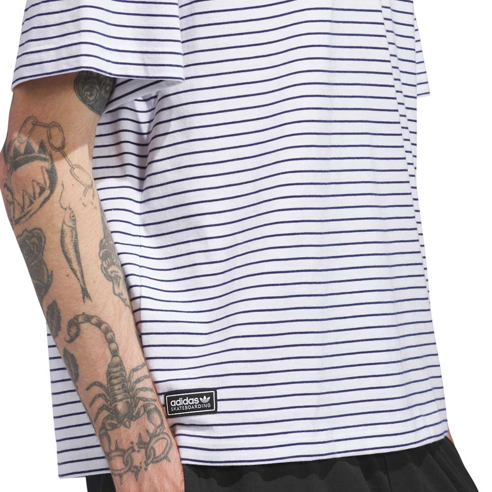 A person wearing the Skate Yarn Dye Tee White Dark Blue (KC6291) from Adidas Skateboarding, a white t-shirt with thin dark blue horizontal stripes and an adidas Skateboarding label, shows off a tattooed arm featuring a scorpion and other designs.