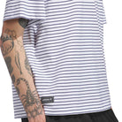 A person wearing the Skate Yarn Dye Tee White Dark Blue (KC6291) from Adidas Skateboarding, a white t-shirt with thin dark blue horizontal stripes and an adidas Skateboarding label, shows off a tattooed arm featuring a scorpion and other designs.