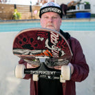 A man in a cap and jacket holds a skateboard vertically, showing its graphic deck and Stage 4 Eric Dressen Pro 151 Black Silver Standard INDY Trucks Set of TWO. A swimming pool and plants blur in the background.