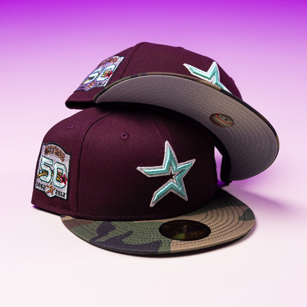 Two Swag Era 50th Camo Maroon Houston Astros New Era Hat 5950 Fitted caps, featuring teal star logos, commemorative anniversary patches, and camouflage brims, are stacked on a pink and purple gradient background.