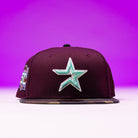 The Swag Era 50th Camo Maroon Houston Astros New Era 5950 Fitted Hat features a maroon design with a teal and white star, a 50th Camo Maroon anniversary side patch, and stands out against a purple gradient background.