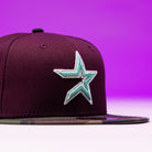 The Swag Era 50th Camo Maroon Houston Astros New Era Hat 5950 Fitted features a teal and white embroidered star logo on the front and a camouflage-patterned flat brim, presented against a purple background.