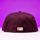 A maroon Swag Era 50th Camo Houston Astros New Era 5950 Fitted Hat is shown from the back, with a small rectangular MLB logo patch near the bottom, set against a vibrant purple background.