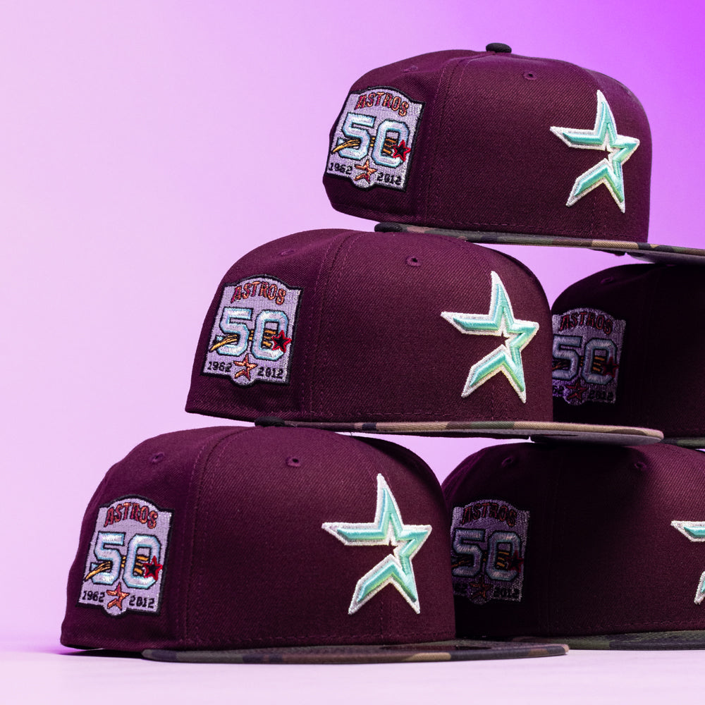 Five Swag Era 50th Camo Maroon Houston Astros New Era Hat 5950 Fitted caps are stacked in a pyramid against a purple gradient background, each featuring a teal star logo and a 50th Anniversary patch.