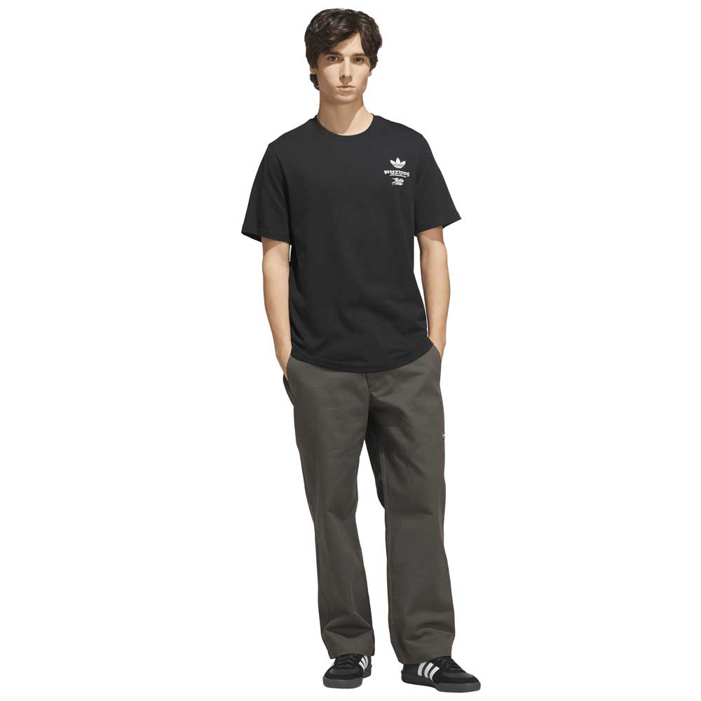 A young man stands with hands in pockets, wearing the black and white TOS Crane Tee (KS4336) from Adidas Skateboarding featuring a white logo, paired with dark gray pants and black Adidas sneakers—an understated look that hints at an adidas collaboration.