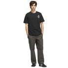 A young man stands with hands in pockets, wearing the black and white TOS Crane Tee (KS4336) from Adidas Skateboarding featuring a white logo, paired with dark gray pants and black Adidas sneakers—an understated look that hints at an adidas collaboration.