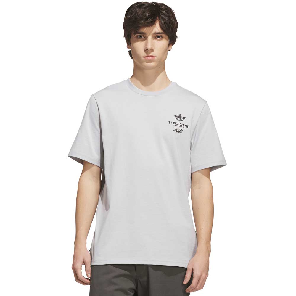A young man with dark hair stands against a plain white background, wearing the Adidas Skateboarding TOS Crane Tee Gray KY7718—a light gray short-sleeve t-shirt featuring a small black graphic and “Temple of Skate” text on the left chest.