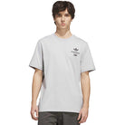 A young man with dark hair stands against a plain white background, wearing the Adidas Skateboarding TOS Crane Tee Gray KY7718—a light gray short-sleeve t-shirt featuring a small black graphic and “Temple of Skate” text on the left chest.