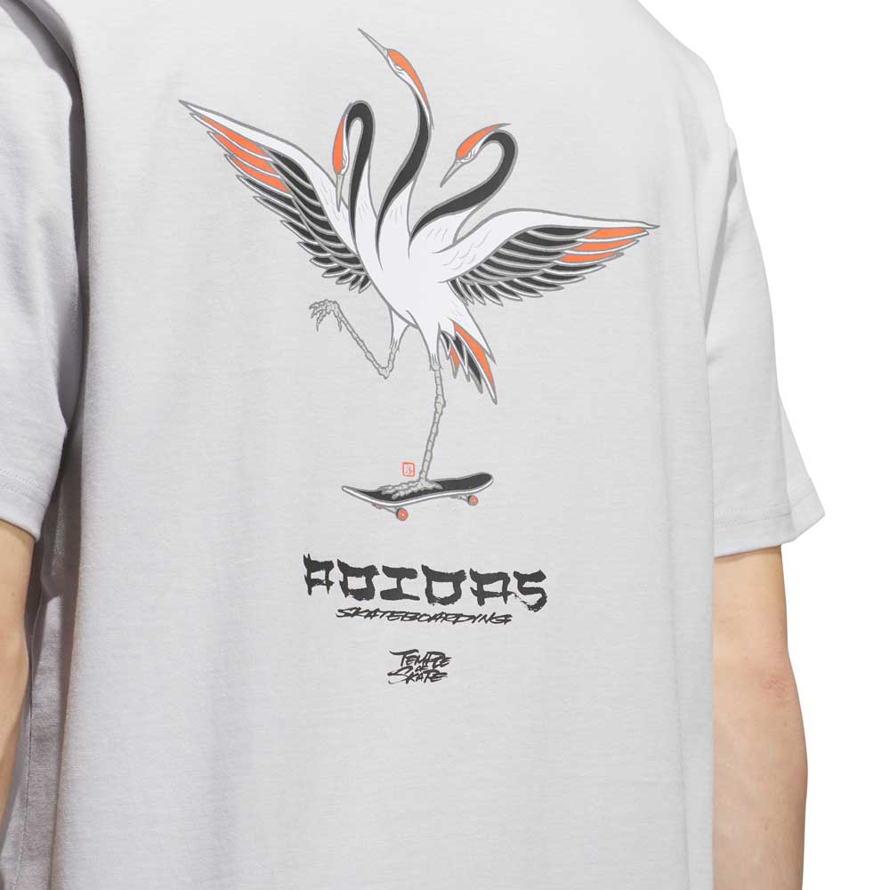 A person wearing the TOS Crane Tee Gray (KY7718) from Adidas Skateboarding—a skateboarding t-shirt featuring an illustration of two cranes on a skateboard with stylized text and signatures below the image, inspired by the Temple of Skate collaboration.