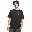 A young man with brown hair wears the TOS Curb Tee Black Multi KS4338 from Adidas Skateboarding—a plain black t-shirt featuring three small adidas Originals trefoil logos on the left chest—and stands against a white background, capturing the essence of skate culture.