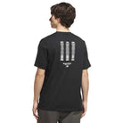 A person wearing the adidas Skateboarding TOS Curb Tee Black Multi (style KS4338), featuring three vertical, patterned stripes and small white text on the back. The individual stands with their back to the camera, capturing classic skate culture style.