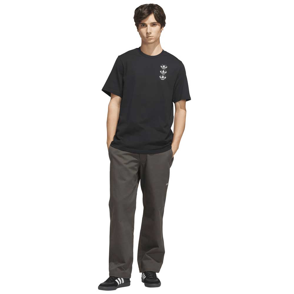 A young man stands with his hands in his pockets, wearing the TOS Curb Tee Black Multi KS4338 from Adidas Skateboarding—featuring small white graphics on the chest—paired with dark gray pants and black adidas Originals sneakers with white stripes. He faces the camera against a white background, showcasing a look inspired by modern skate culture.