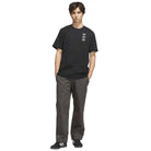 A young man stands with his hands in his pockets, wearing the TOS Curb Tee Black Multi KS4338 from Adidas Skateboarding—featuring small white graphics on the chest—paired with dark gray pants and black adidas Originals sneakers with white stripes. He faces the camera against a white background, showcasing a look inspired by modern skate culture.
