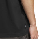 A close-up of a person’s arm and hand resting by their side, wearing the TOS Curb Tee Black Multi KS4338 from Adidas Skateboarding, featuring a small black tag on the seam, paired with dark pants. The white background reflects influences from skate culture.
