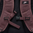 Close-up of the maroon shoulder straps on the Tattoo Black Light Violet FD7544502 Nike SB RPM Skate Backpack, featuring a black Nike logo patch and center buckle fastener, set against a black background.