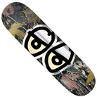 The Team Eyes Camo Foil 8.5 Krooked Skateboards Deck features a camo foil pattern of leaves and bark, accented with bold cartoon-style eyes with black outlines and cream centers in the center.