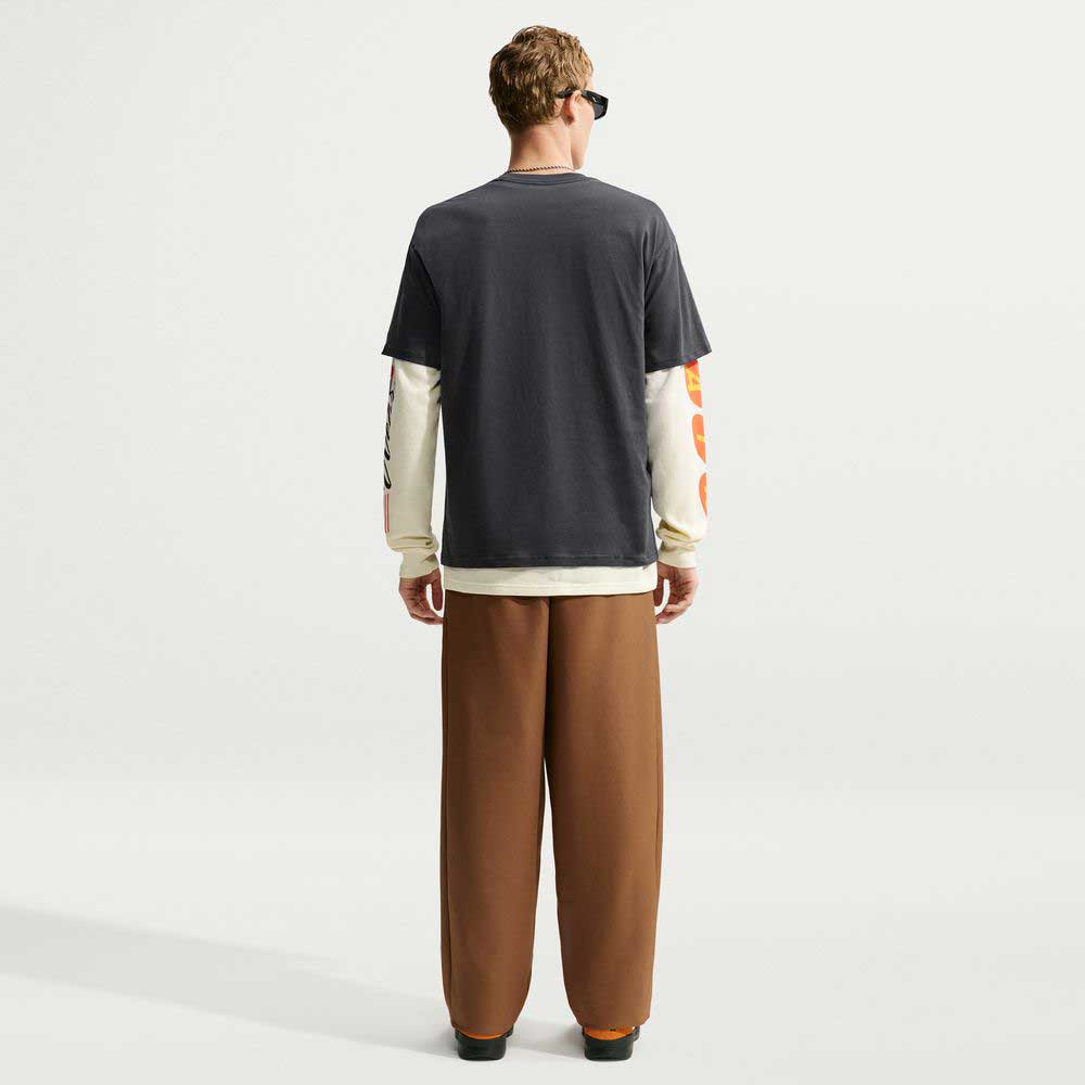 Back view of a person wearing a Tee Black IW3134060 T-Shirt Nike SB Skateboarding over long cream-colored sleeves with orange graphics on the arms and brown trousers.