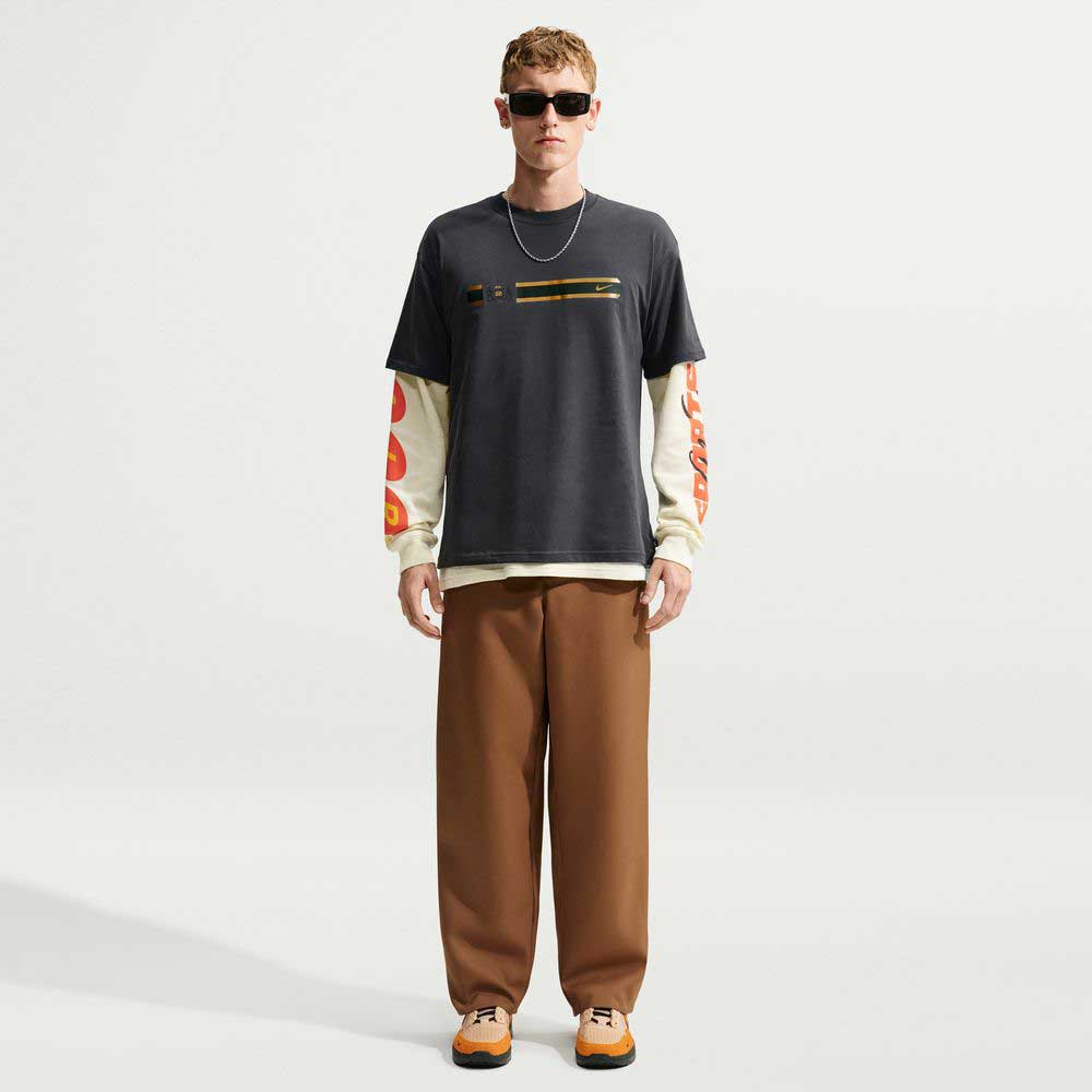 A man wearing a black Nike SB Skateboarding Tee Black IW3134060 T-Shirt over a cream-colored long-sleeved shirt with graphic prints on the sleeves, paired with brown trousers and sneakers.