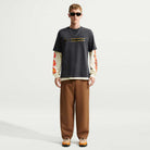 A man wearing a black Nike SB Skateboarding Tee Black IW3134060 T-Shirt over a cream-colored long-sleeved shirt with graphic prints on the sleeves, paired with brown trousers and sneakers.