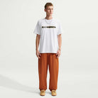 A person wearing a Tee White IW3134100 T-Shirt Nike SB Skateboarding with a horizontal black stripe across the chest, a crest, and a small logo, paired with loose-fitting rust-colored pants.