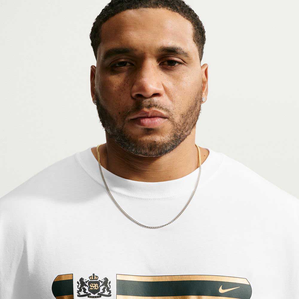 A man wearing a white Nike SB Tee White IW3134100 T-Shirt Nike SB Skateboarding featuring a gold and black crest graphic on the chest.