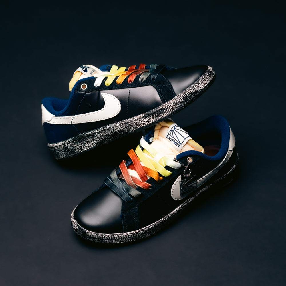 Two blue Zoom Tennis Classic x Rassvet IO9846400 Nike SB leather skateboarding shoes with sail-colored swooshes are set against a dark background. Each sneaker features rainbow-colored gradient laces arranged differently, while the comfortable insole ensures all-day support both on and off the board.