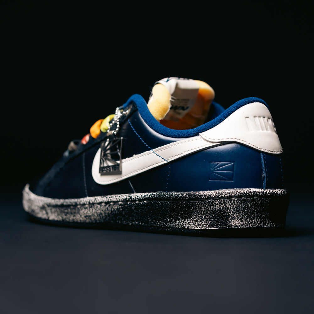 A pair of Zoom Tennis Classic x Rassvet IO9846400 Blue Void Sail Nike SB leather skateboarding shoes with a navy blue and white upper, unique patterns, yellow accents, and a speckled black-and-white sole is displayed against a dark background. Designed as premium skate shoes, they also feature a cushioned Solarsoft insole for enhanced comfort.