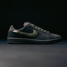The Zoom Tennis Classic HF7386200 in Deep Pewter and Medium Olive Suede, from Nike SB Skateboarding, takes inspiration from skate shoe heritage, featuring a dark metallic swoosh logo, a textured sole, and matching laces. Designed with a supportive Solarsoft insole for comfort, this black sneaker is showcased against a dark, minimal background.