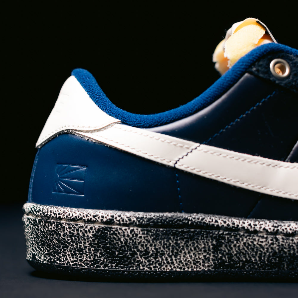 Close-up of a Nike SB Zoom Tennis Classic x Rassvet IO9846400 skateboarding shoe in Blue Void Sail, featuring a navy blue leather upper, white stripe, blue suede accents, white heel tab, and a speckled black-and-white sole against a black background. Designed for skateboarding comfort with a supportive insole.