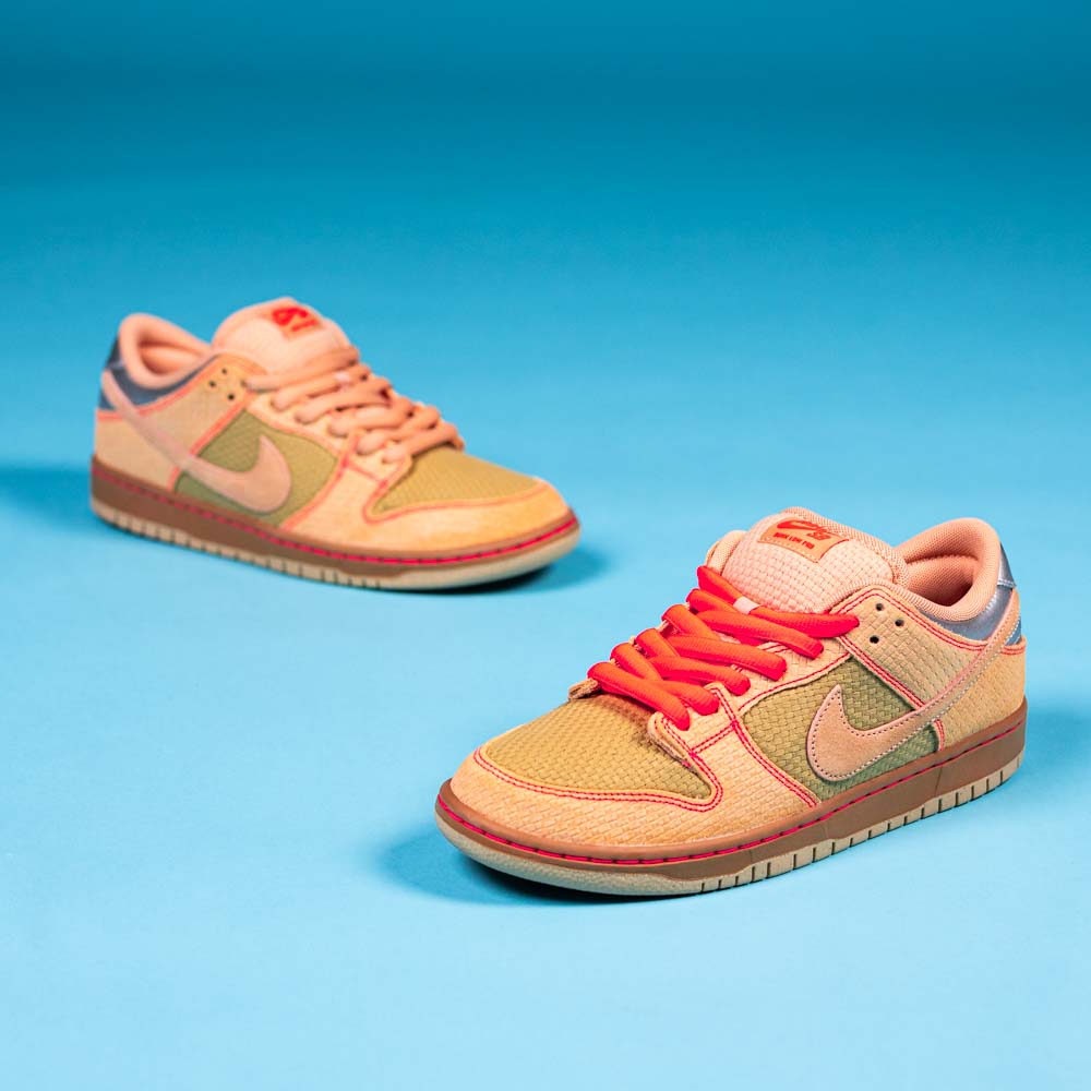 A pair of READ BELOW the BUTTON RAFFLE Nike SB Som Tum Dunk Low sneakers with orange, beige, and pink accents sits on a bright blue surface against a matching blue background.