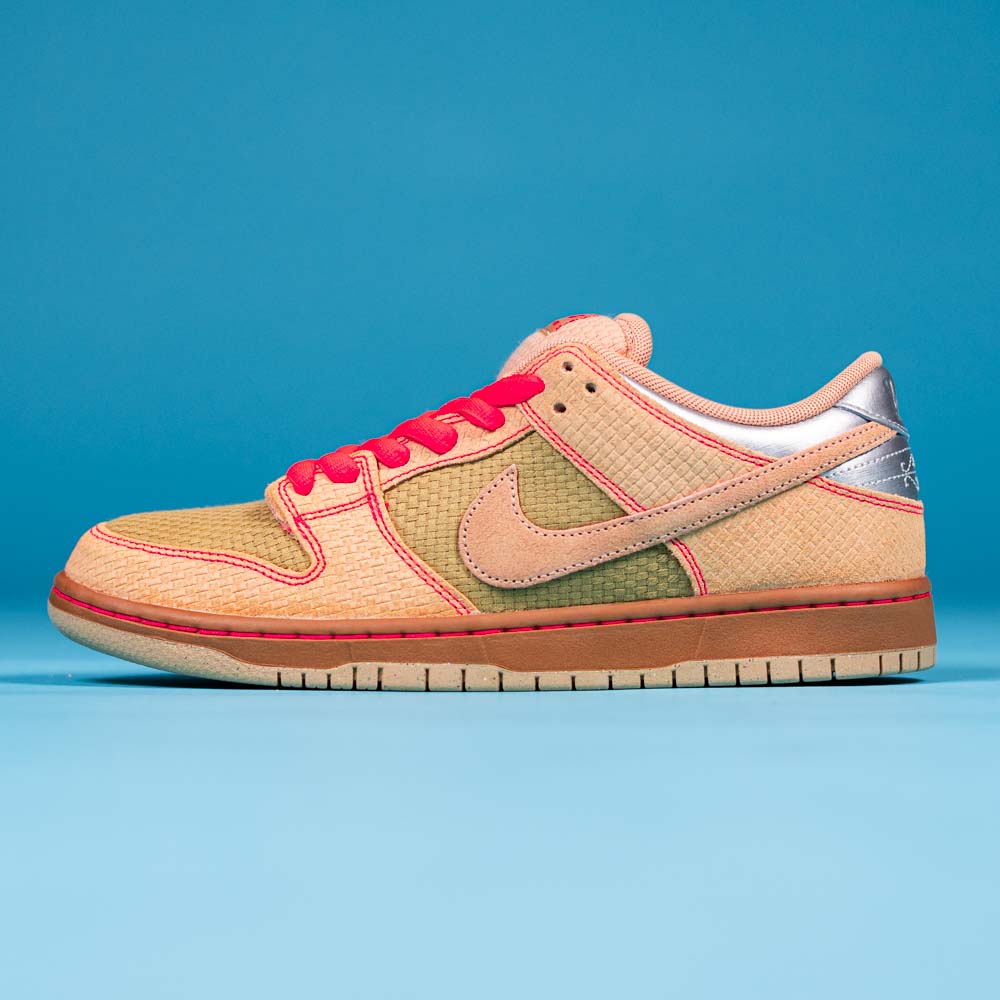 The READ BELOW the BUTTON RAFFLE Nike SB Som Tum Dunk Low, featuring a textured tan and green upper with pink accents and silver heel detail, is displayed on a blue background—enter the raffle at Southside Skatepark Skateshop.