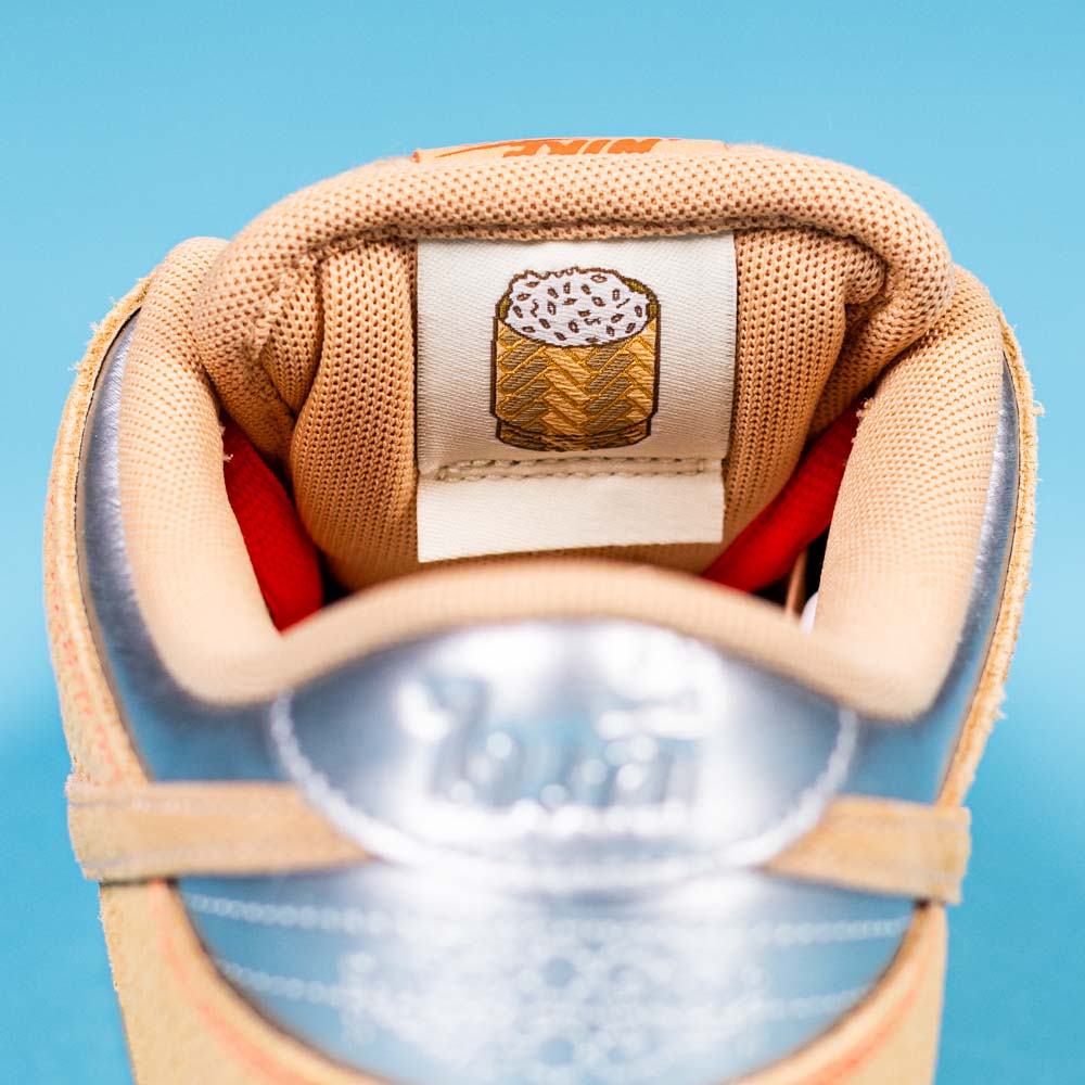 Close-up of the tan and silver READ BELOW the BUTTON RAFFLE Nike SB Som Tum Dunk Low sneaker tongue with a burrito tag on a light blue background—a must-have for any sneaker raffle or Southside Skatepark Skateshop fan.