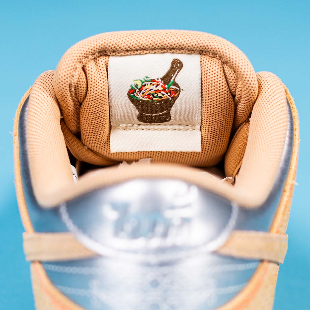 Close-up of a tan READ BELOW the BUTTON RAFFLE Nike SB Som Tum Dunk Low tongue, featuring an embroidered mortar and pestle patch with colorful ingredients, set against a blue background for the Southside Skatepark Skateshop raffle.