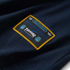 A close-up of navy fabric displays a rectangular patch with Adidas branding, yellow border, blue and white text, and small logos—stitched onto the Thrasher Magazine x Adidas AFA Tee Navy KX4543 Skateboarding shirt.