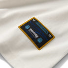 A close-up of the Thrasher Magazine x Adidas AFA Tee White KX4548, featuring a white fabric with a rectangular black patch displaying blue and white text, gold borders, and Adidas logos—a limited-edition Skateboarding collab label.