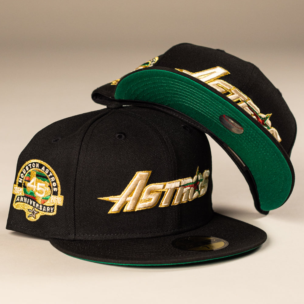 The La Morenita Black 45th Houston Astros New Era 5950 Fitted hat features gold Astros lettering, a green underbill, and a 45th anniversary patch; one stands upright while another snapback rests on top.